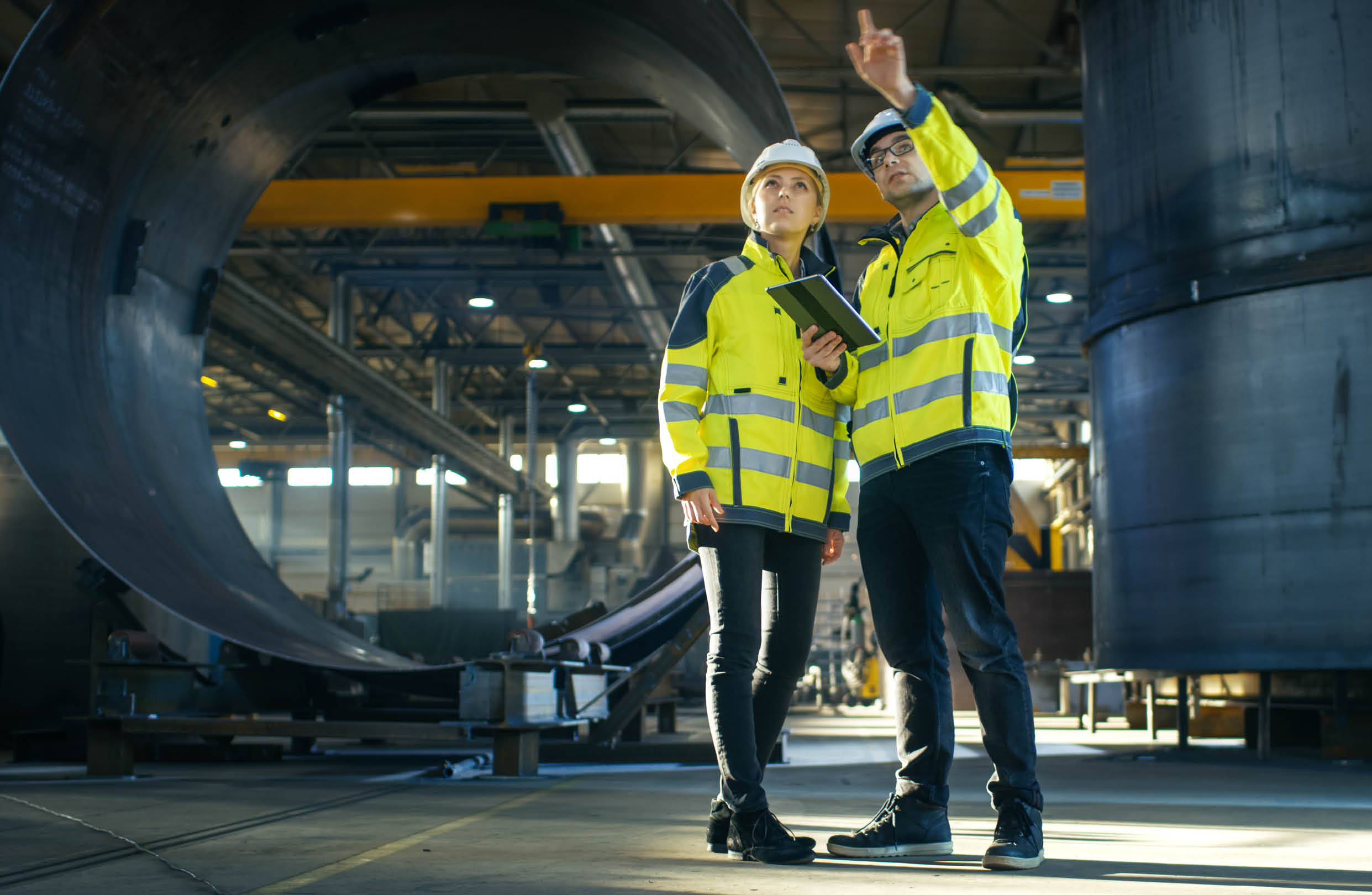 Male and Female Industrial Engineers in Hard Hats Discuss New Project while Using Tablet Computer. They're Making Calculated Engineering Decisions.They Work at the Heavy Industry Manufacturing Factory.