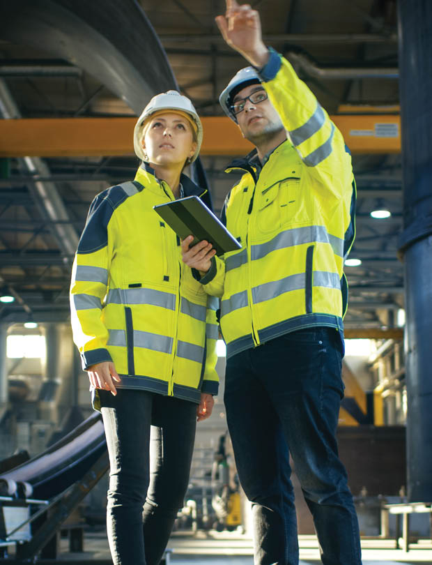 Male and Female Industrial Engineers in Hard Hats Discuss New Project while Using Tablet Computer. They're Making Calculated Engineering Decisions.They Work at the Heavy Industry Manufacturing Factory.